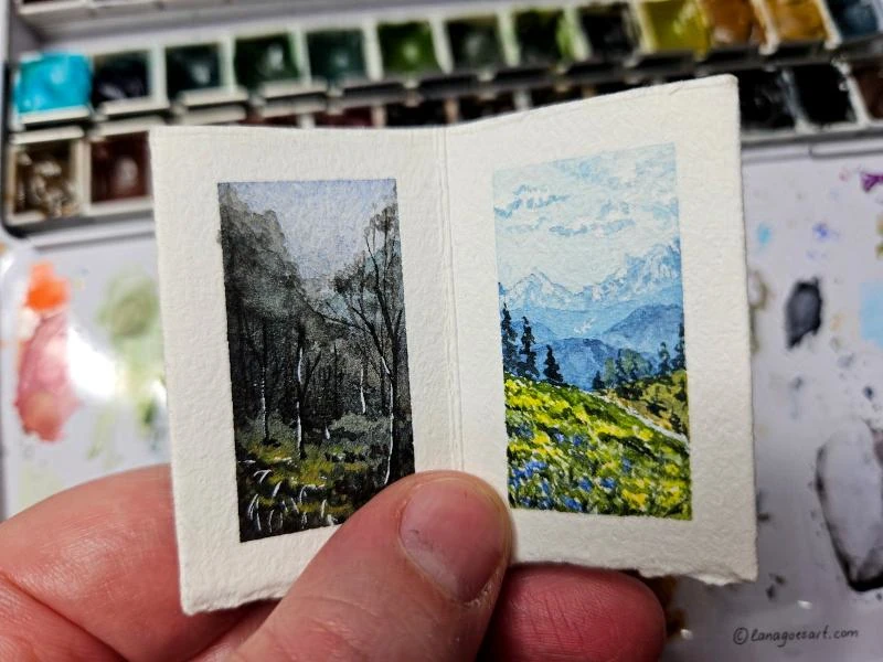 Tiny sketchbook held in a hand with two open pages showing landscapes painted in watercolor.