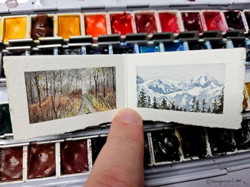 Tiny sketchbook with two open pages in landscape format showing a forest and a mountain landscape painted in watercolors.