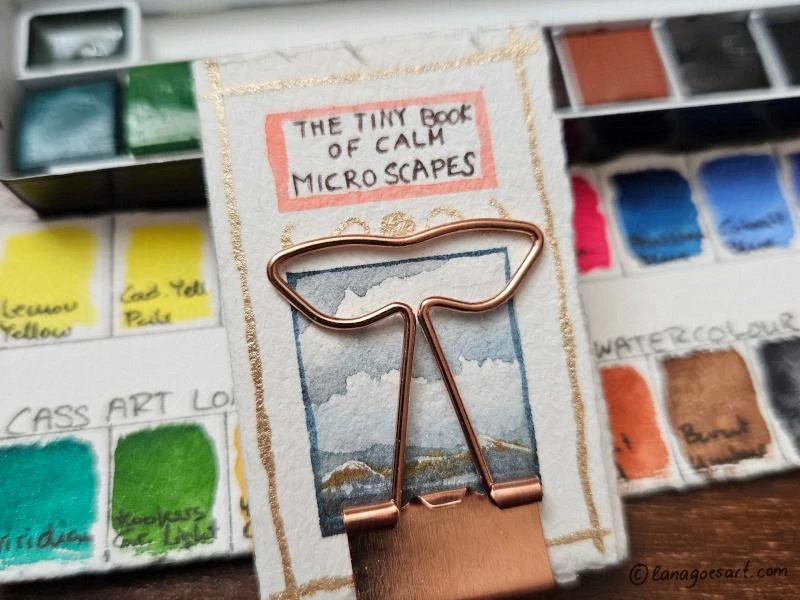 Tiny sketchbook held closed by a clip with a small palette in the background.